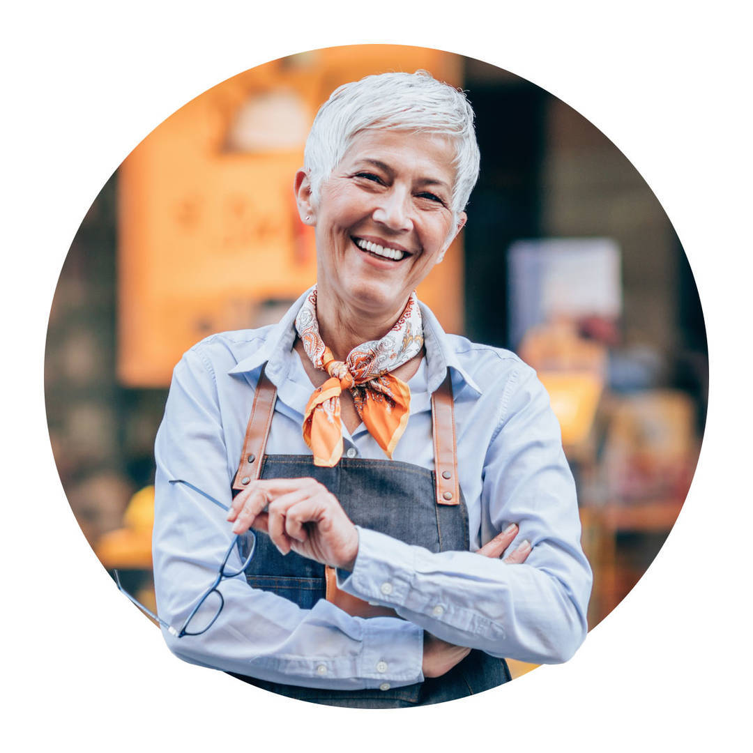 Mature female business owner, with short white hair, proudly standing in front of shop with arms crossed.